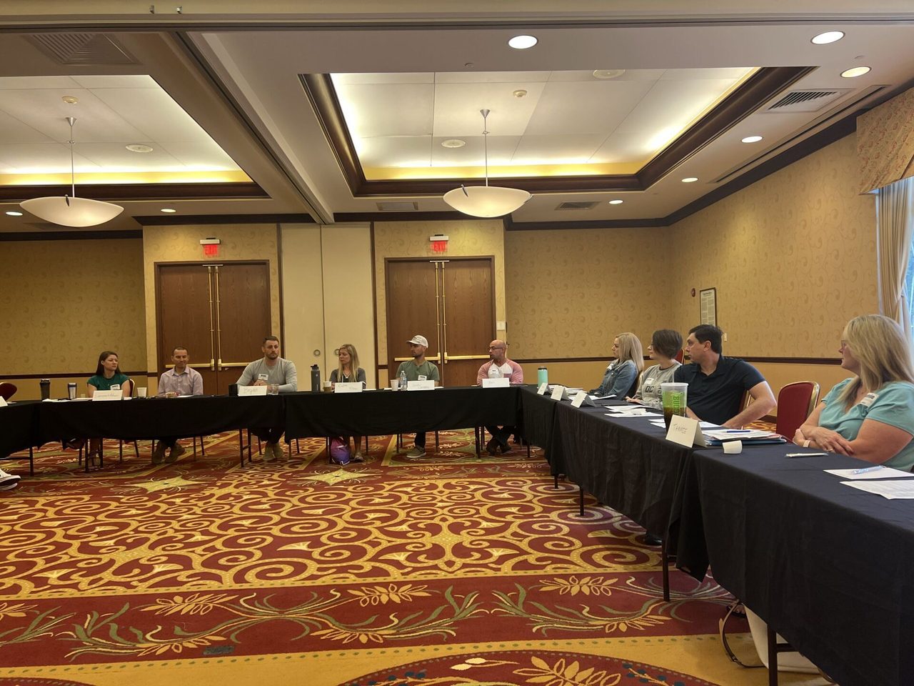 A group of people are sitting at long tables in a conference room.