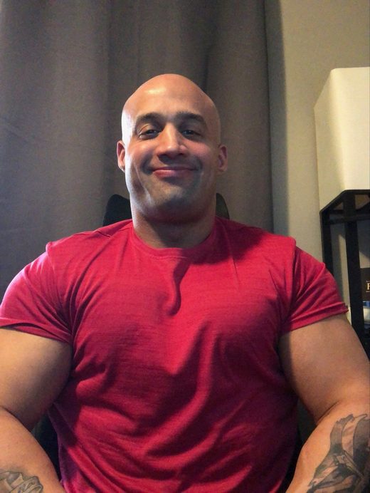 A man with a tattoo on his arm is wearing a red shirt and smiling.