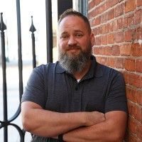 A man with a beard and a dark polo shirt standing in front of a brick wall.