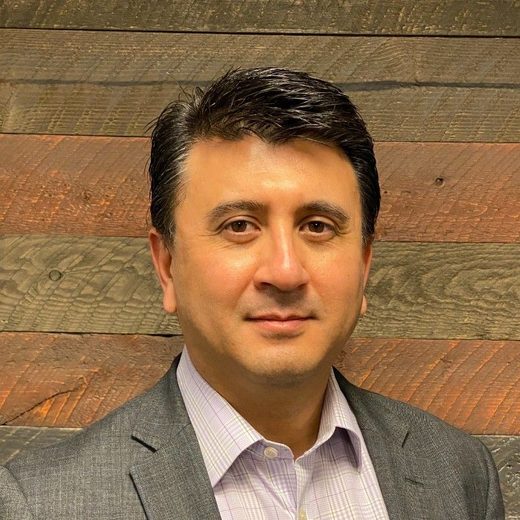 A man in a grey jacket and pink shirt in front of a wooden wall.