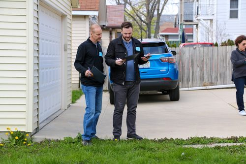 Two men are standing in front of a house looking at a tablet.