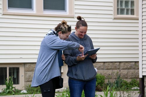 Two women are standing in front of a house looking at a tablet.