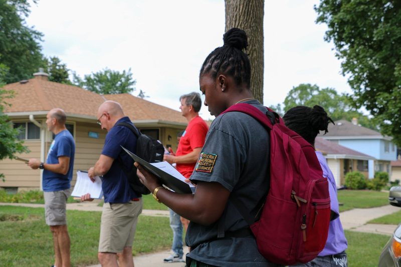 A group of people are standing outside of a house looking at a clipboard.