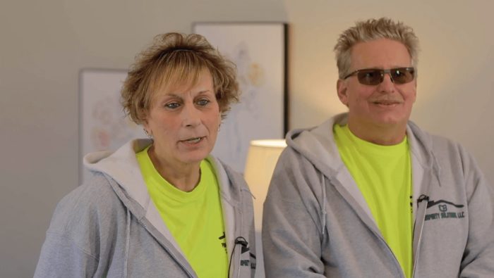 A man and a woman wearing hoodies and neon yellow shirts are standing next to each other in a living room.